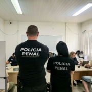 Foto mostra dois policiais penais, de costa, com camisetas pretas da Polícia Penal, na frente de uma sala onde atendentes estão coletando os dados dos detentos.