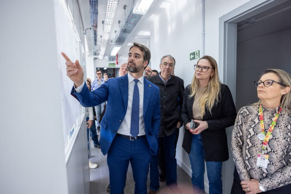 Corredor interno de um edifício recém-estruturado, onde um grupo acompanha uma vistoria técnica; uma pessoa aponta para informações fixadas na parede enquanto as demais observam, em ambiente bem iluminado com acabamento moderno.