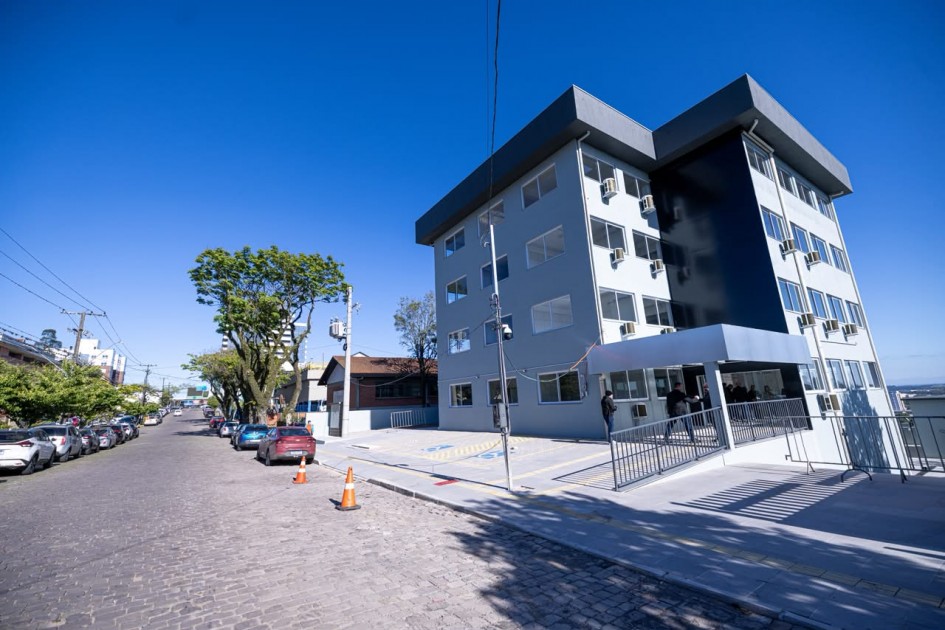 Vista externa de um prédio institucional de vários andares, com fachada em tons de cinza e azul, acesso por rampa e estacionamento frontal. A construção está localizada em uma rua arborizada com veículos estacionados ao longo da via.