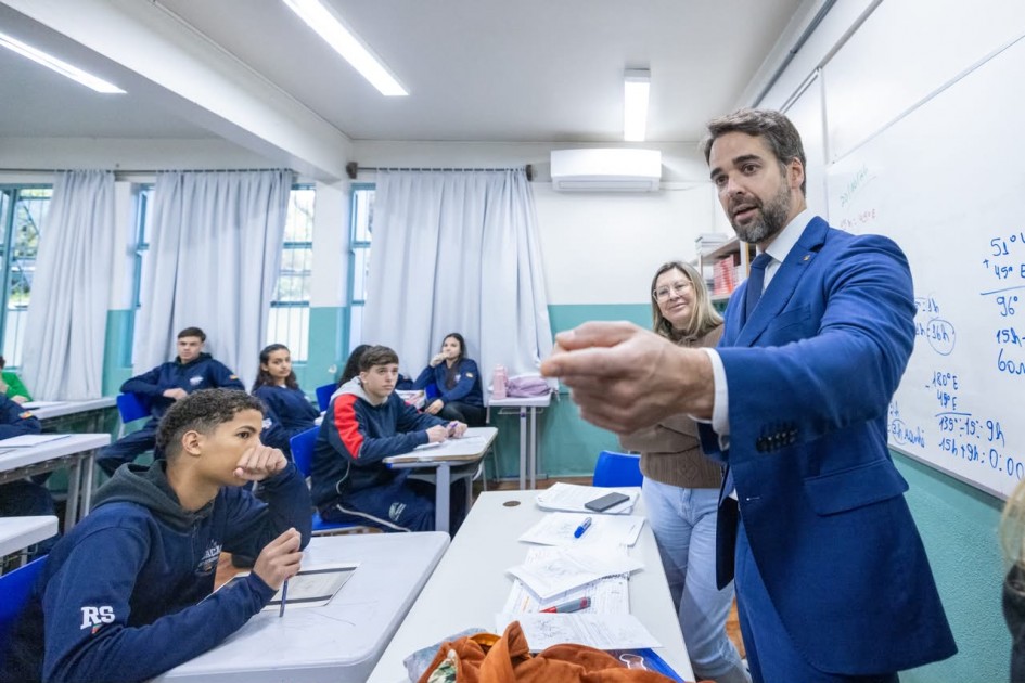 Sala de aula com estudantes sentados em carteiras enquanto o governador fala junto ao quadro branco. O espaço possui iluminação clara, cortinas nas janelas e materiais escolares sobre a mesa do professor.