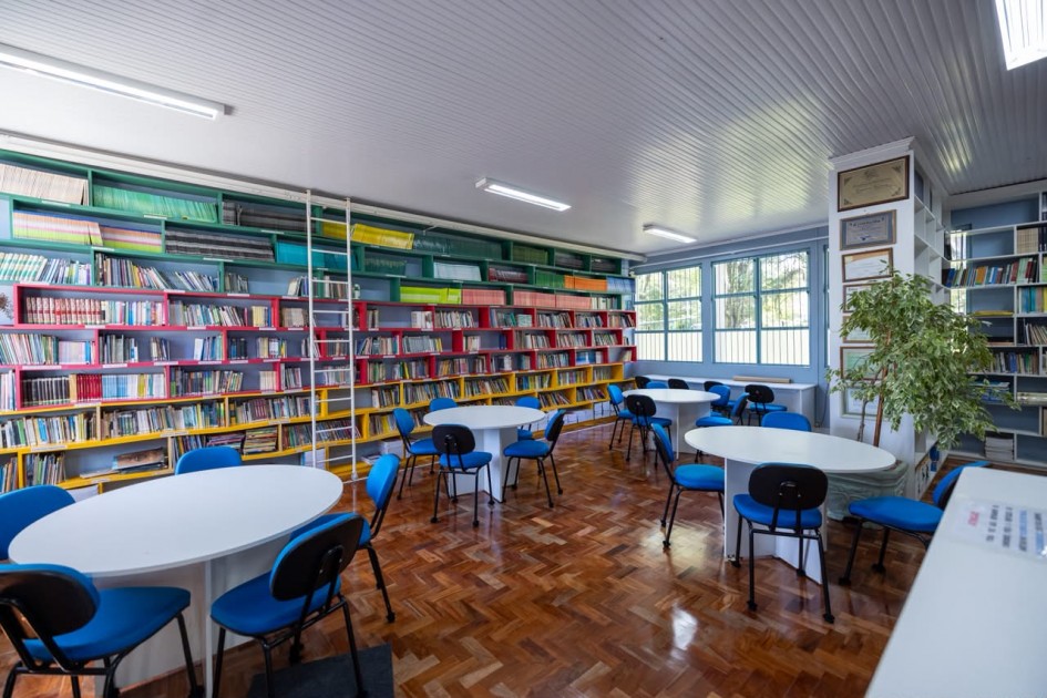 Biblioteca escolar com estantes coloridas repletas de livros, mesas redondas e cadeiras azuis distribuídas pelo espaço. O ambiente é amplo, bem iluminado e organizado para leitura e estudos.
