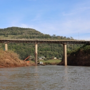 Foto mostra o rio com a ponte ao fundo. Atrás, aparece um morro verde. Na margem esquerda, há raízes expostas, provavelmente arrastadas pela enchente.