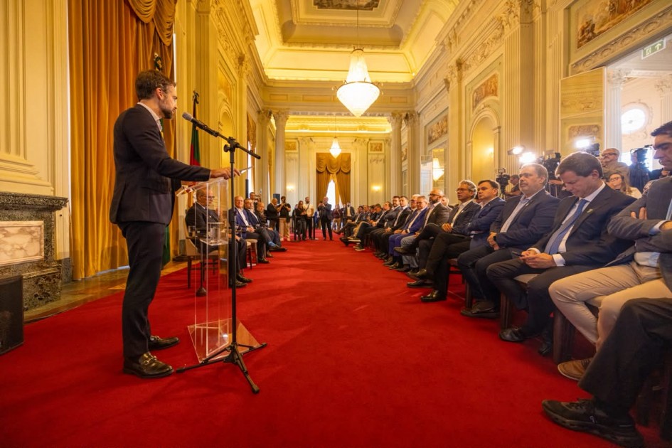 Cerimônia em salão histórico com tapete vermelho, onde uma pessoa faz um pronunciamento em púlpito transparente diante de uma plateia de autoridades e convidados. O ambiente possui arquitetura clássica, grandes lustres e cobertura da imprensa.