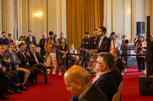 Evento institucional em salão nobre, com uma pessoa discursando ao microfone enquanto participantes e profissionais de comunicação acompanham a apresentação. O espaço tem decoração clássica, cortinas douradas e iluminação interna destacada.