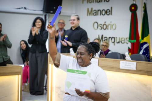 Foto mostra uma formanda, uma mulher negra de camiseta branca, levantando o canudo de formatura bem alto e mostrando o diploma. Ela ergue o braço bem alto com o diploma e sorri muito. Ao fundo, pessoas aplaudem a cena.