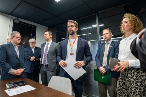 Uma fotografia colorida em plano médio-geral e perspectiva em contra-plongée suave (vista de ângulo levemente baixo) registra o governador Eduardo Leite acompanhado por secretários e autoridades durante anúncio institucional sobre a estruturação de fundo de investimento para apoio a empresas do RS. **As Autoridades e o Governador (ao centro e em primeiro plano):** * No centro da composição, o governador Eduardo Leite surge em pé de frente, ligeiramente voltado para a esquerda, segurando papéis/documentos oficiais em ambas as mãos. Ele veste blazer azul-marinho sobre camisola branca de gola redonda, traz fita/cordão institucional ao pescoço com crachá e exibe expressão atenta. * À sua esquerda (à direita do observador), encontram-se dois gestores em pé: * Um homem de blazer azul-escuro, camisa social clara, gravata bege e calças de padrão xadrez/médio, segurando uma pasta institucional verde do Banrisul sob o braço. * Uma mulher de cabelos encaracolados volumosos em tom castanho-claro/dourado, vestindo camisa social branca de mangas compridas e saia/calça com estampa de leopardo/onça, segurando um pequeno bloco de notas/agenda. * À sua direita (à esquerda do observador), alinham-se três autoridades e gestores em pé: * Um homem de fato cinzento e gravata roxa/lilás com traços em xadrez. * Um gestor de cabelos grisalhos e fato azul-marinho de braços cruzados ao fundo. * Na extremidade esquerda, um homem de cabelos e barbas grisalhas, óculos e fato escuro com as mãos entrelaçadas à frente do corpo. **O Ambiente e a Mesa (em primeiro plano e ao fundo):** * Em primeiro plano, do lado esquerdo inferior, sobressai o canto de uma mesa de reuniões em madeira escura com folhas de papel, caneta e um smartphone pousados. Ao lado da mesa, vê-se o encosto de uma cadeira de escritório cinzento-clara. * Ao fundo, o espaço conta com divisórias modernas de vidro fosco com persianas pretas integradas e caixilharia metálica escura. * O teto apresenta acabamento escuro/preto com luminárias retangulares encastradas, proporcionando uma iluminação interior focado e de tom frio.